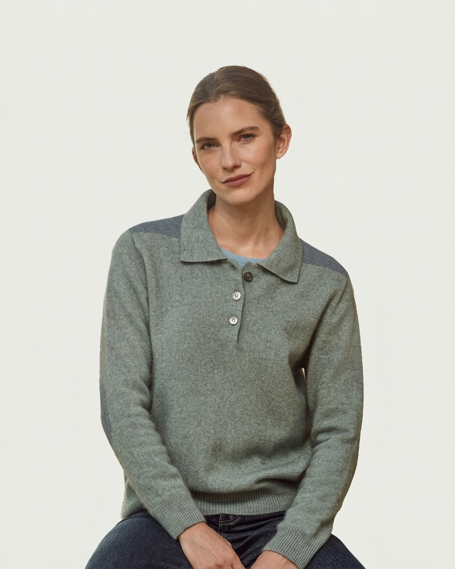 A woman wearing a muted sage green possum merino knit polo sweater with contrasting dark gray shoulder panels, seated against a plain light gray background.