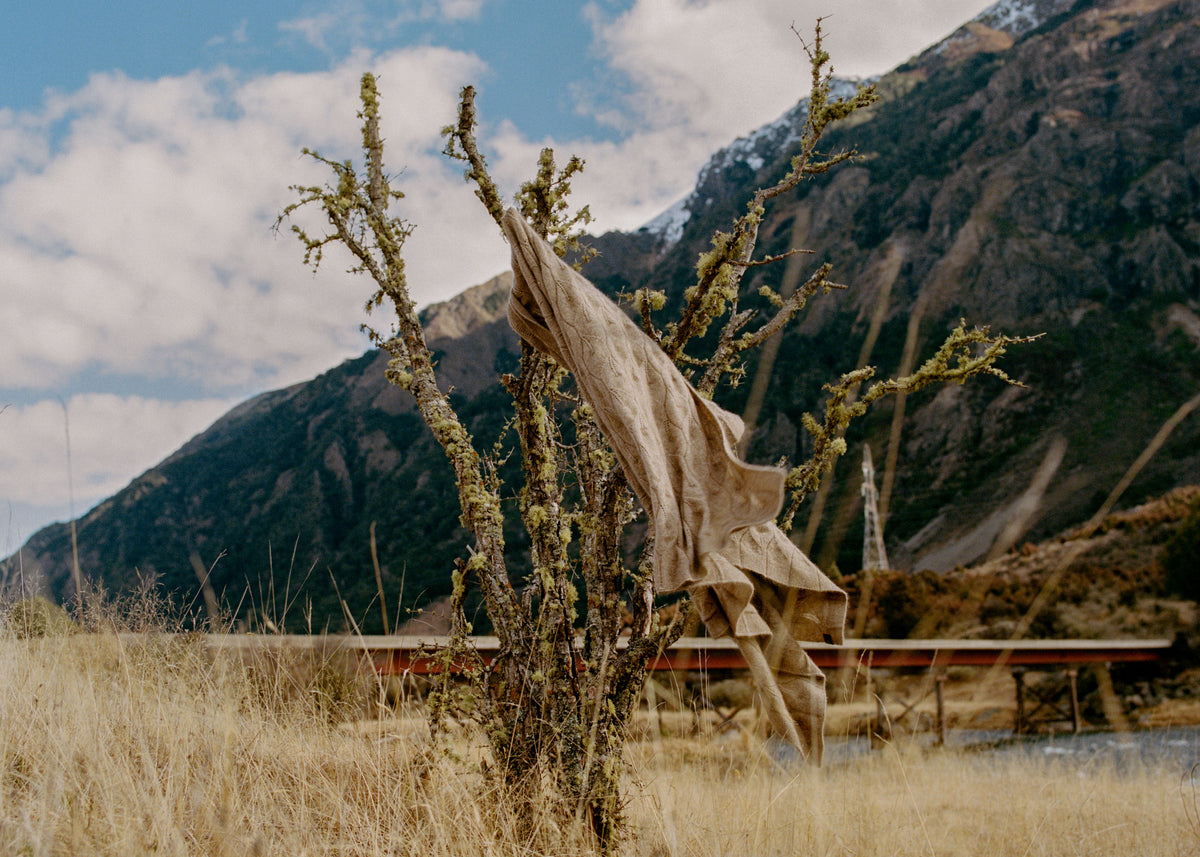 A large beige fabric sculpture draped over a leafless tree in a grassy field, with mountains and a cloudy sky in the background.