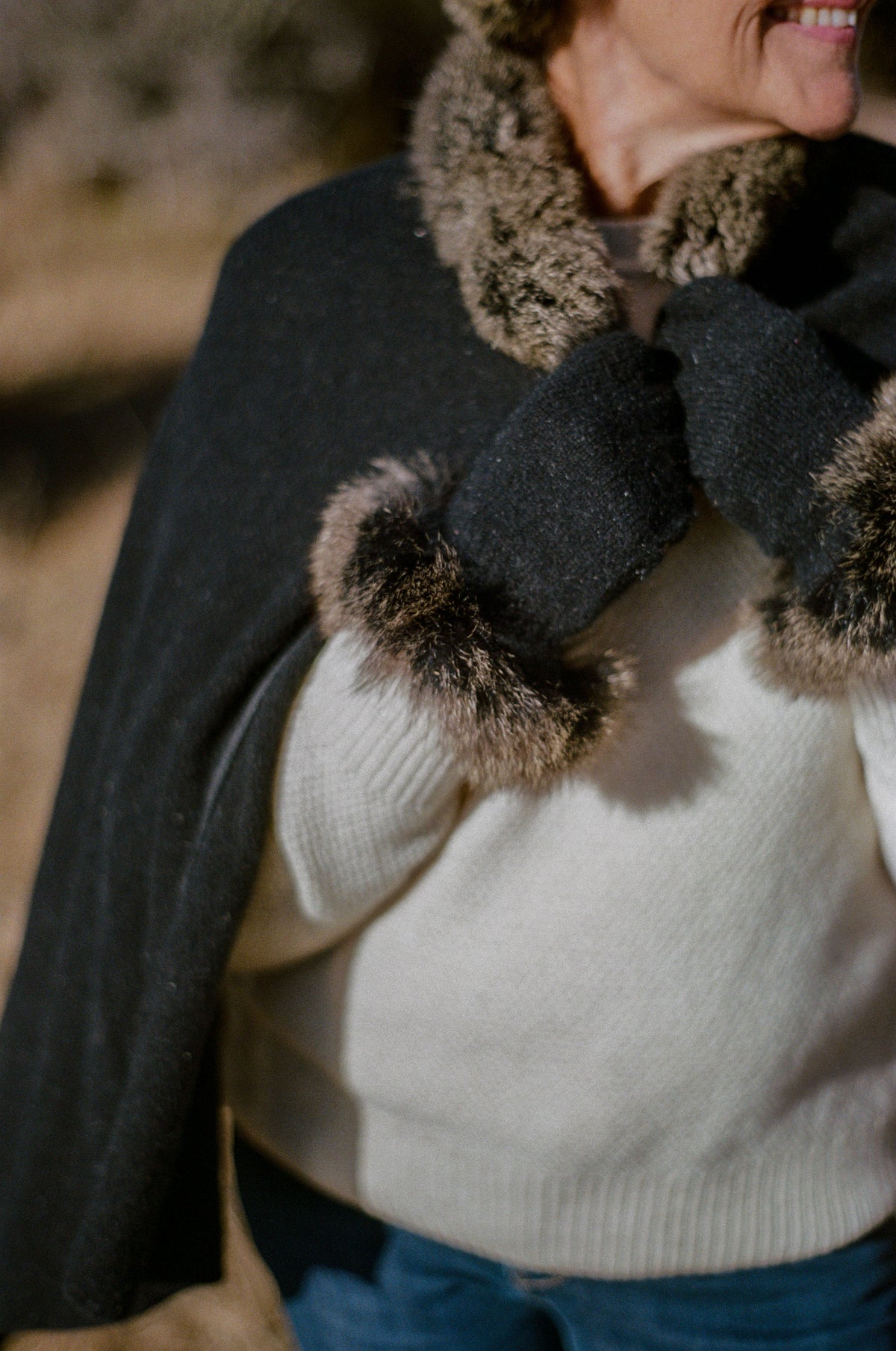 A close-up lifestyle shot of a person wearing a dark charcoal gray cape with a brown and beige faux-fur trim, a cream knit sweater, and blue jeans in an outdoor setting.
