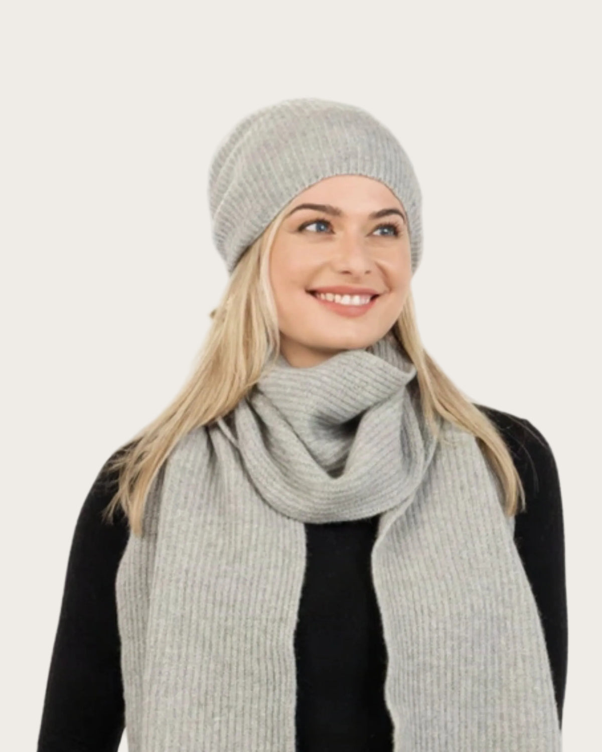 A woman wearing a light gray ribbed knit beanie, matching scarf, and black outfit, photographed against a plain white studio background.
