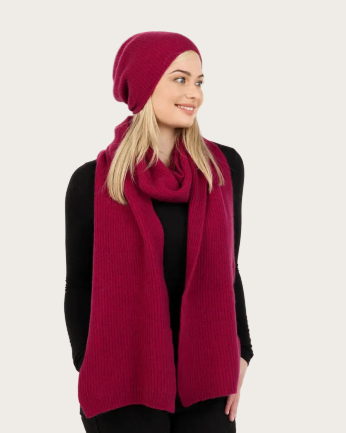 A woman wearing a matching burgundy knit beanie, scarf, and sweater vest over a black top, photographed against a plain light gray studio background.