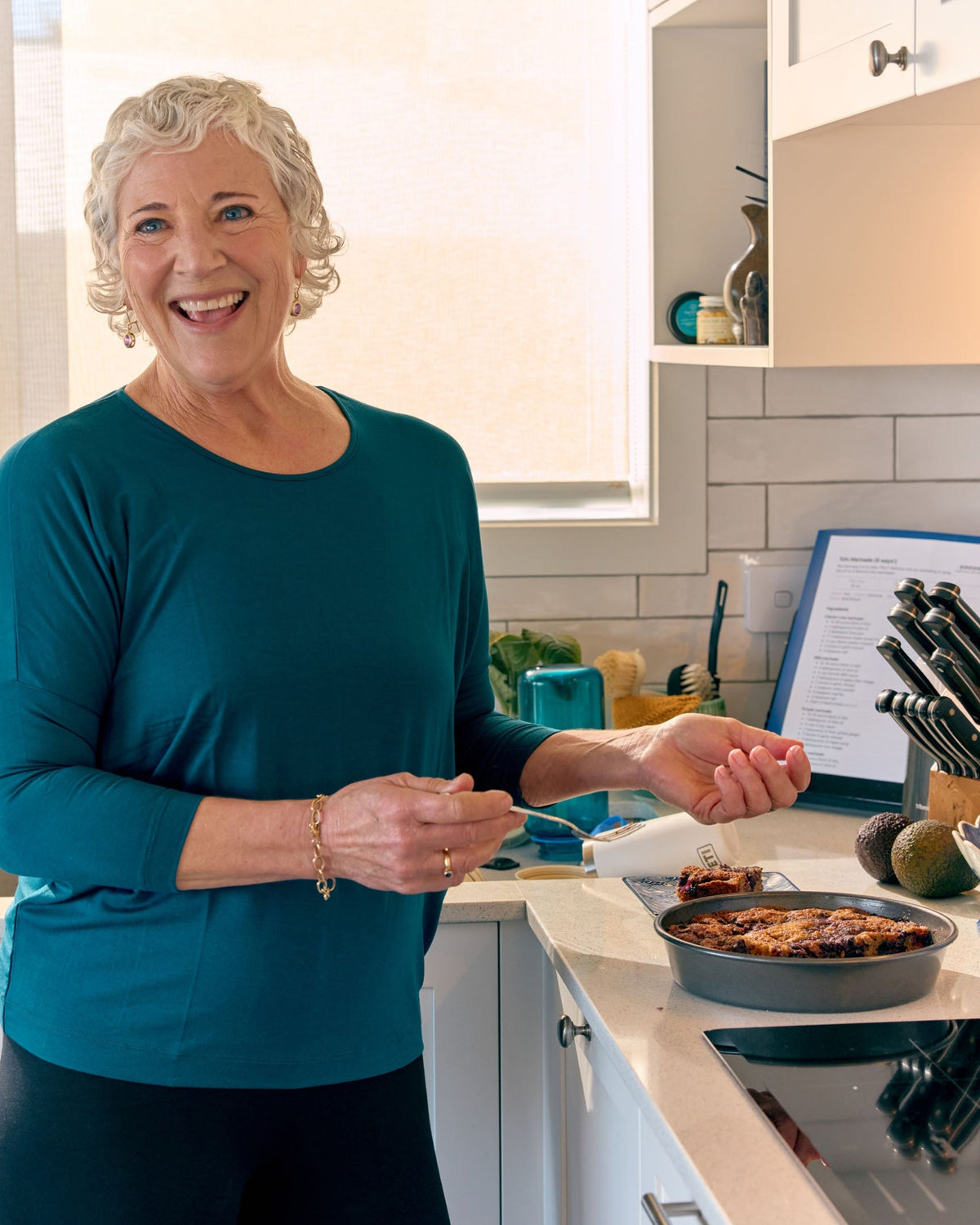 An older woman with short curly gray hair smiling in a bright kitchen, wearing a teal long-sleeve top and black pants. She is standing at a white countertop with a pan of food and a laptop nearby.