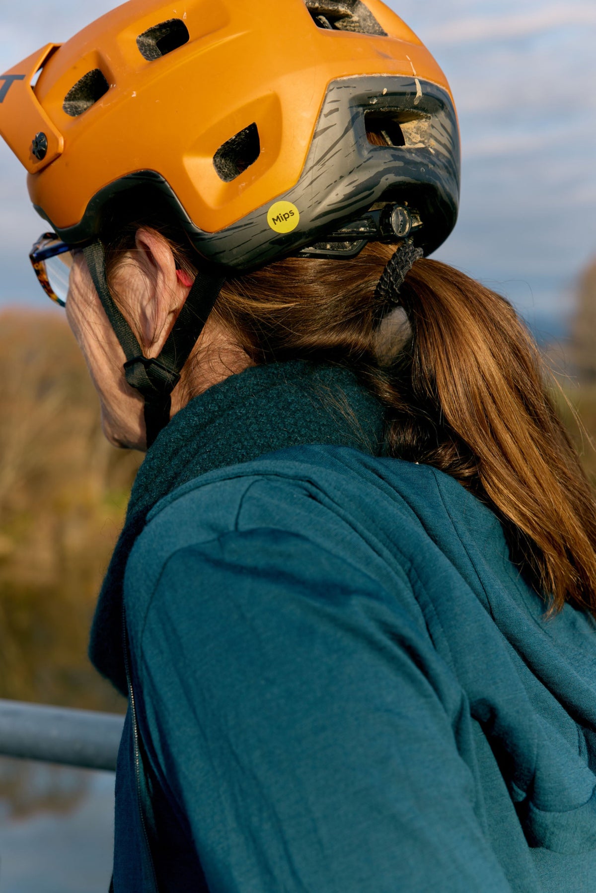 A close-up side profile of a person wearing a bright orange mountain bike helmet with a black chin strap and a teal merino hooded jacket, outdoors with a blurred natural background.