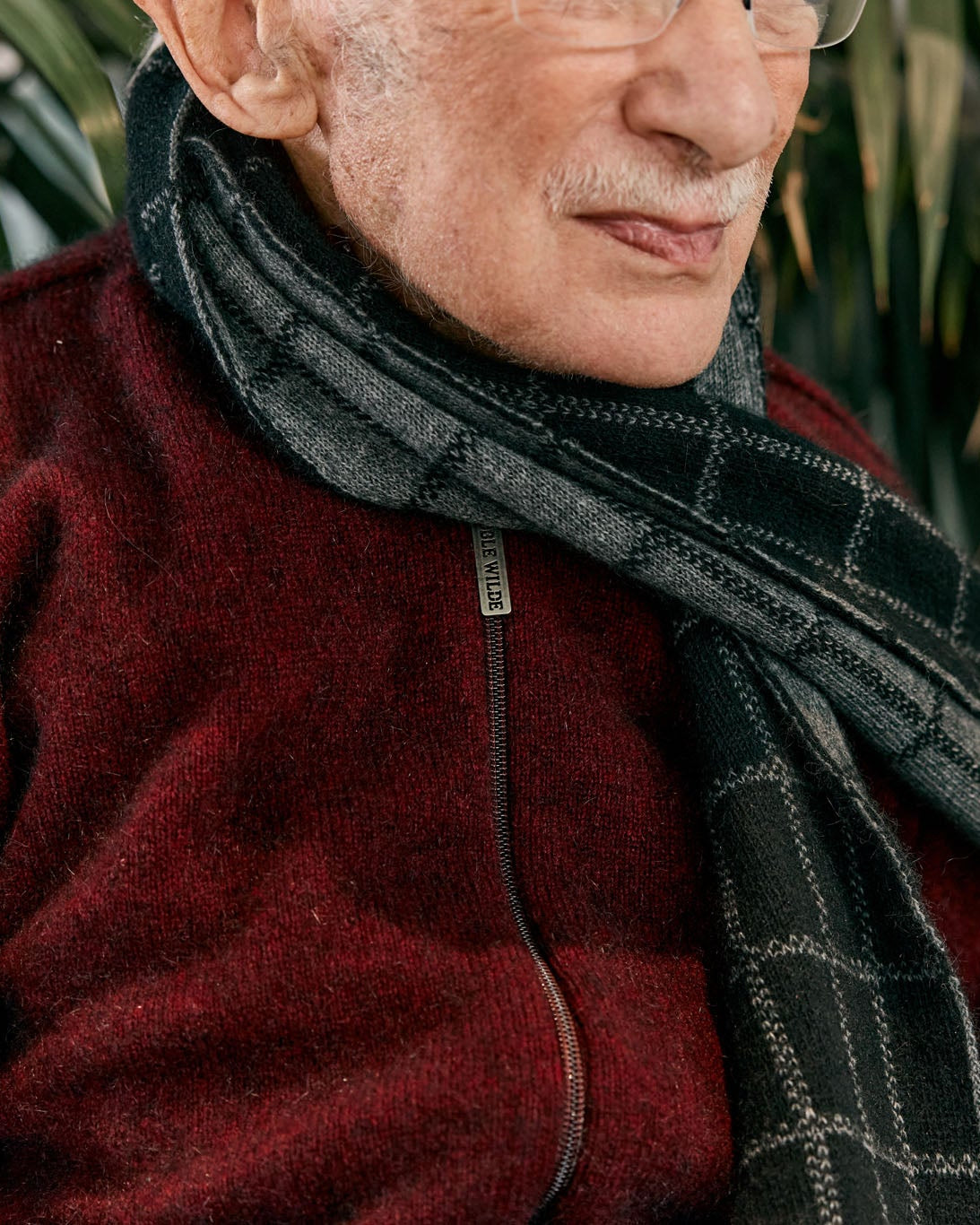 An elderly man wearing a dark red fleece zip-up sweater and a dark gray patterned scarf, with blurred green palm leaves in the background.