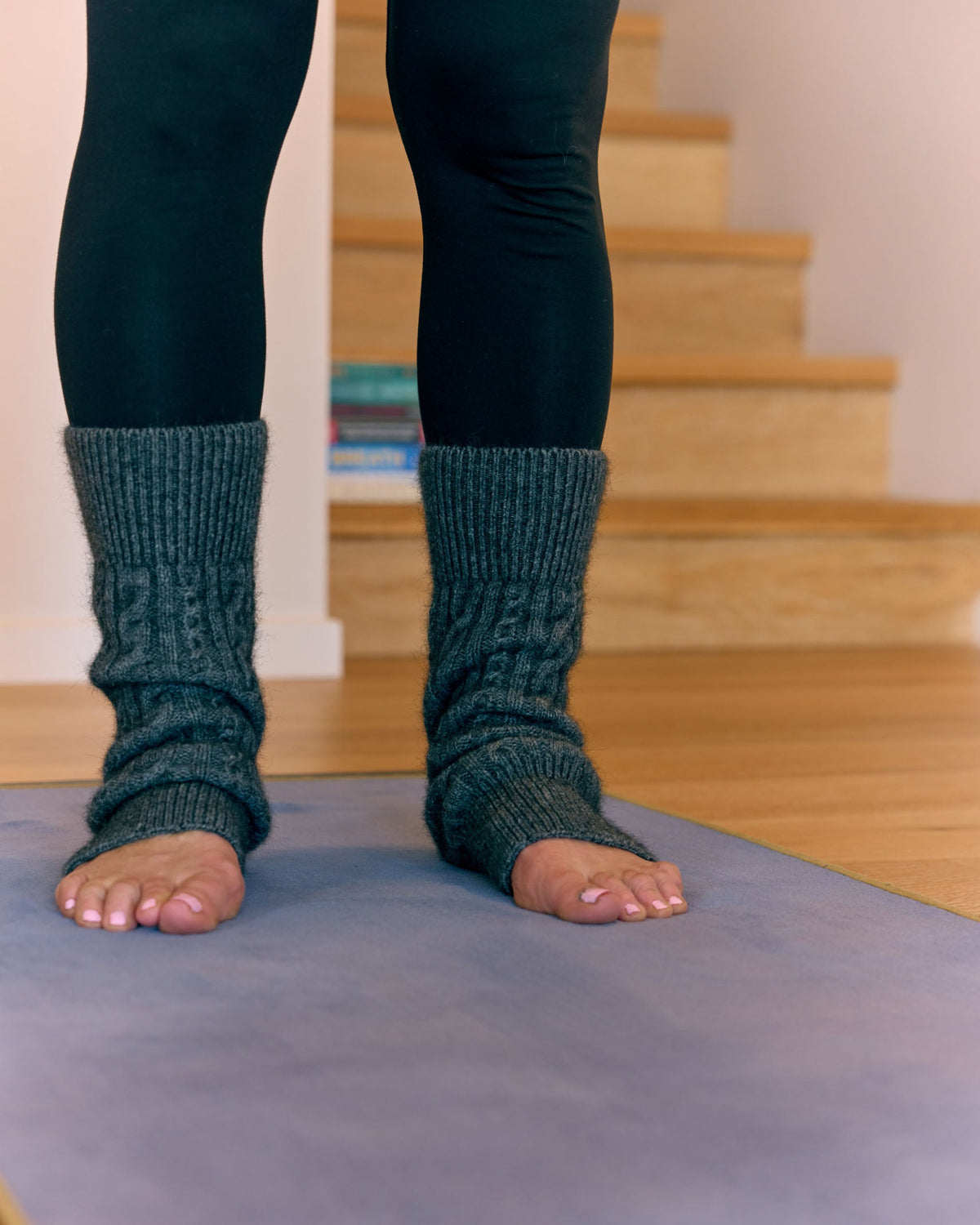 A person wearing dark gray cable-knit leg warmers or foot covers, standing barefoot on a gray yoga mat in front of wooden stairs.