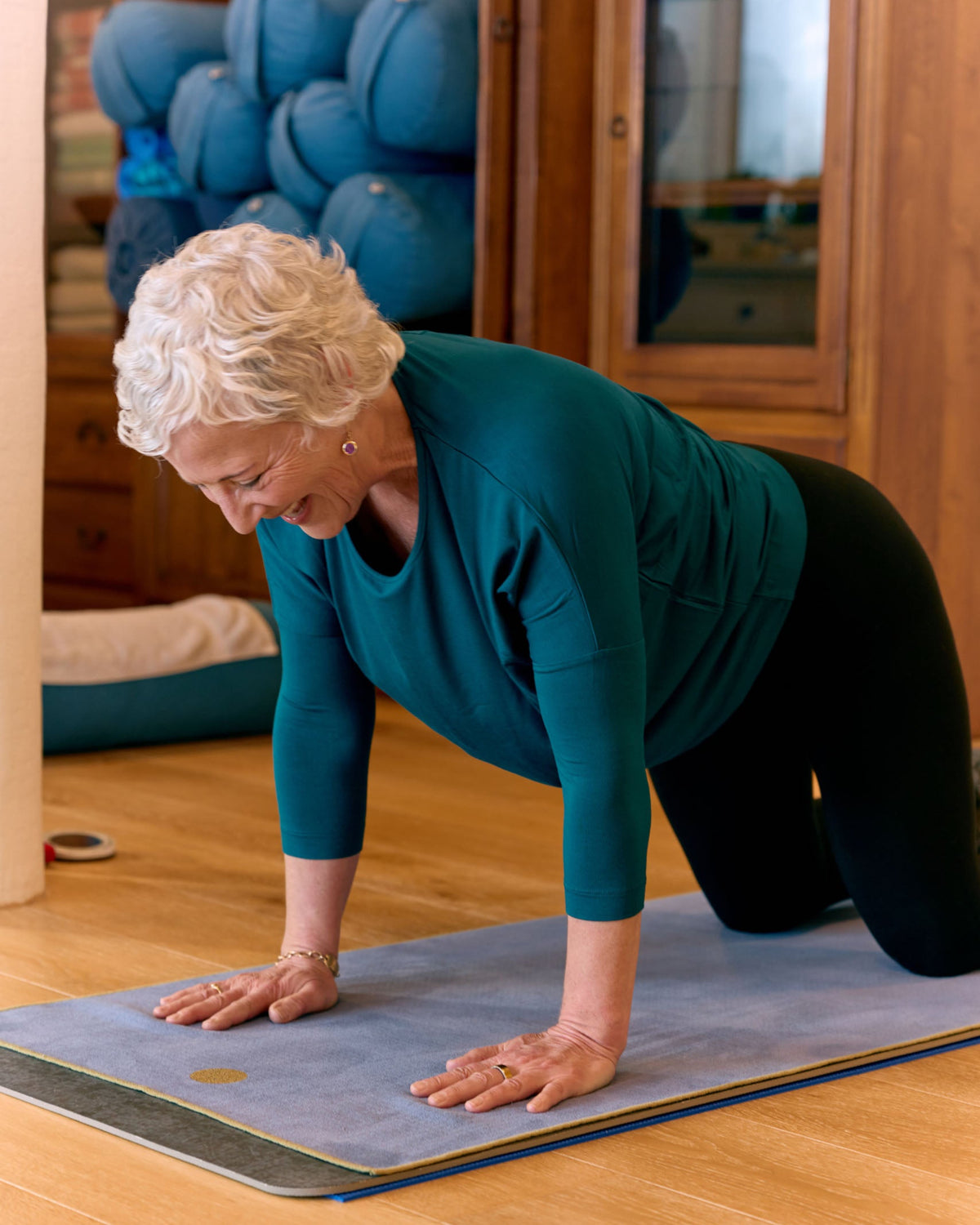 An older woman with short white hair is on a yoga mat in a home studio, wearing a teal long-sleeve top and black leggings while doing a tabletop pose on a wooden floor.