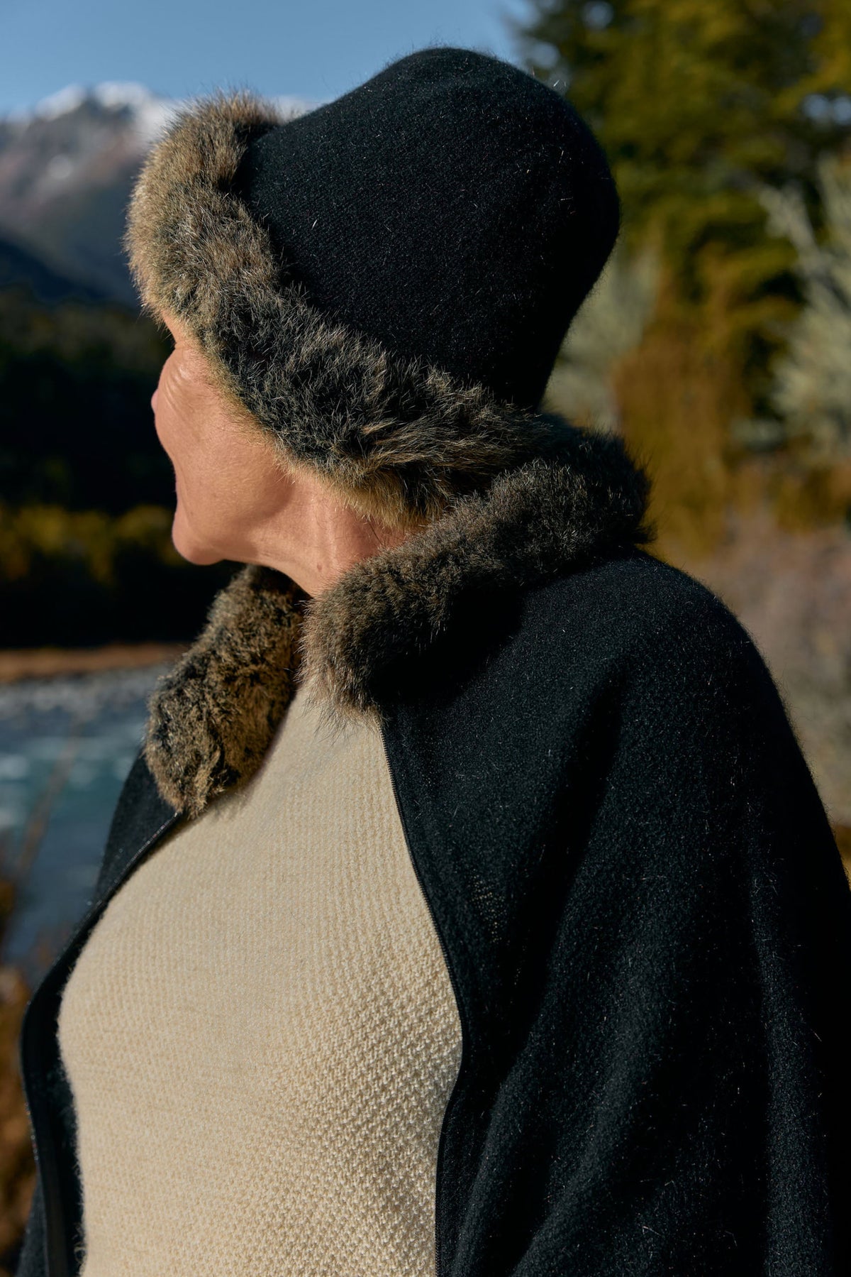 A person shown in side profile wearing a black knit hat with brown faux-fur trim, a black outer layer with matching fur trim, and a cream knit top, photographed outdoors near water and trees.
