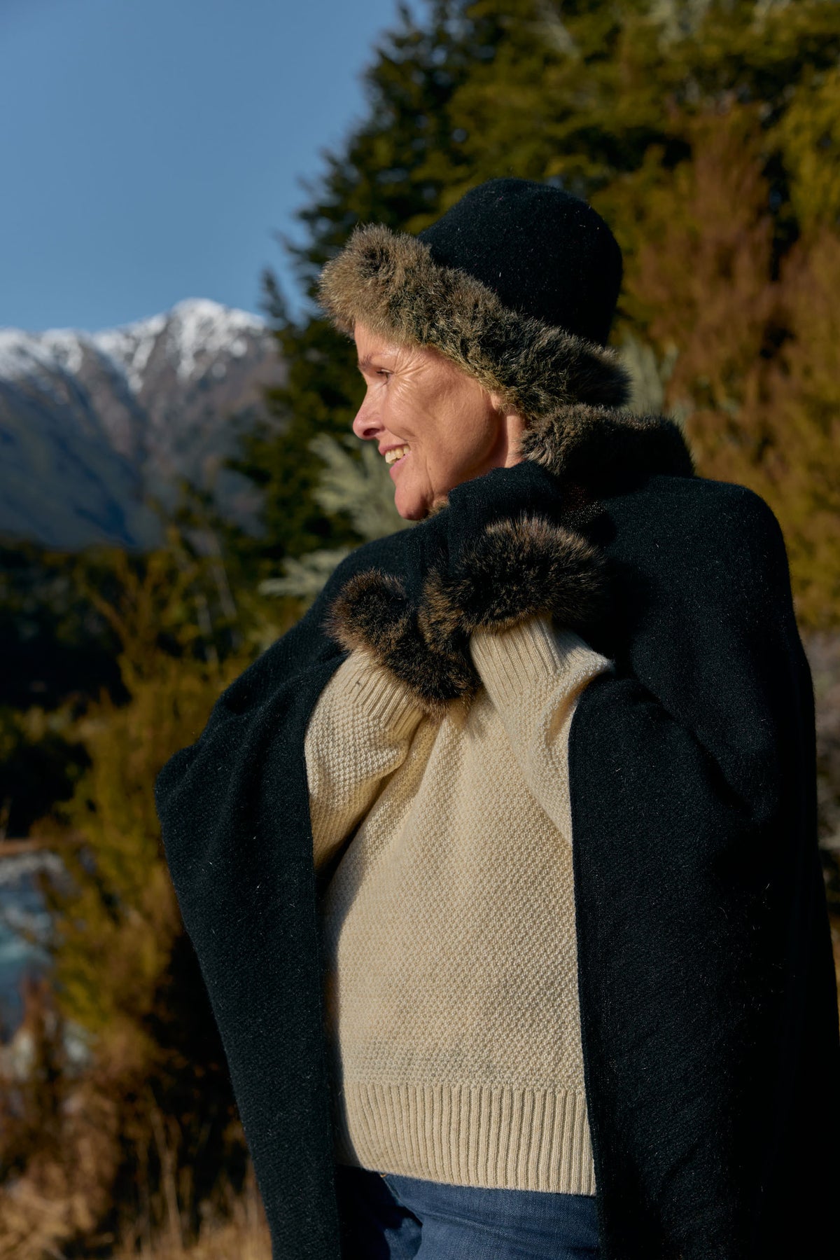 A woman outdoors wearing a black knit sweater, a black coat with dark brown fur trim, a black hat with fur trim, and dark gloves, with mountains and trees in the background.