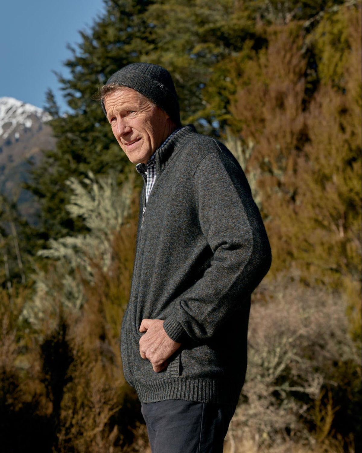 A man wearing a dark gray knit zip-up sweater, dark beanie, and dark trousers stands outdoors in a natural setting with mountains, trees, and shrubs in the background.