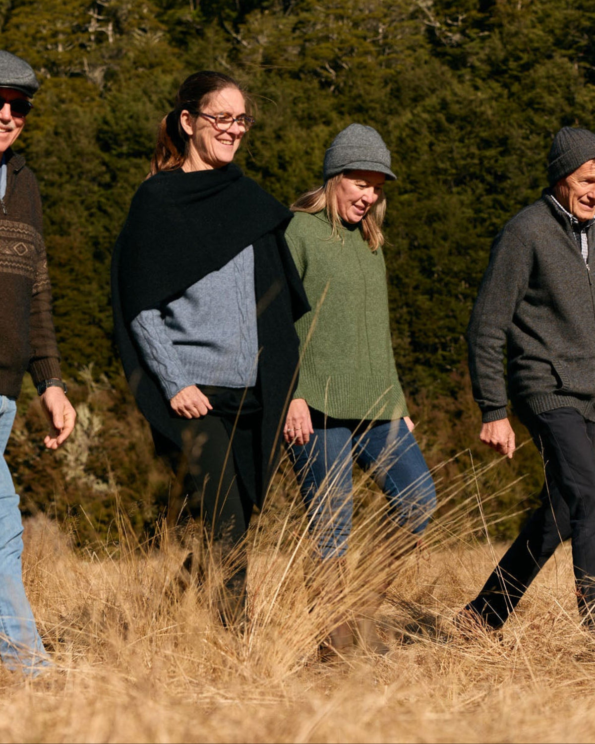 Four people walking through tall dry grass in an outdoor setting with a forest in the background. They are dressed in casual outdoor clothing, including knit sweaters, long sleeves, and beanies.