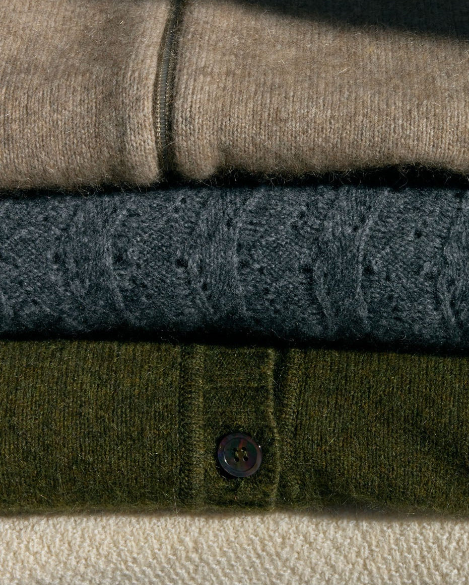 A stack of folded knit sweaters in beige, charcoal gray, olive green, and cream, displayed on a rustic wooden surface against a weathered stone wall.