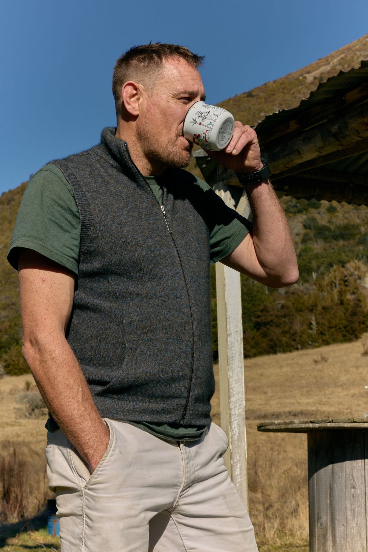 possum merino A man outdoors drinking from a white ceramic mug with a black graphic print, wearing a dark gray zip-up vest over a green t-shirt and beige pants.