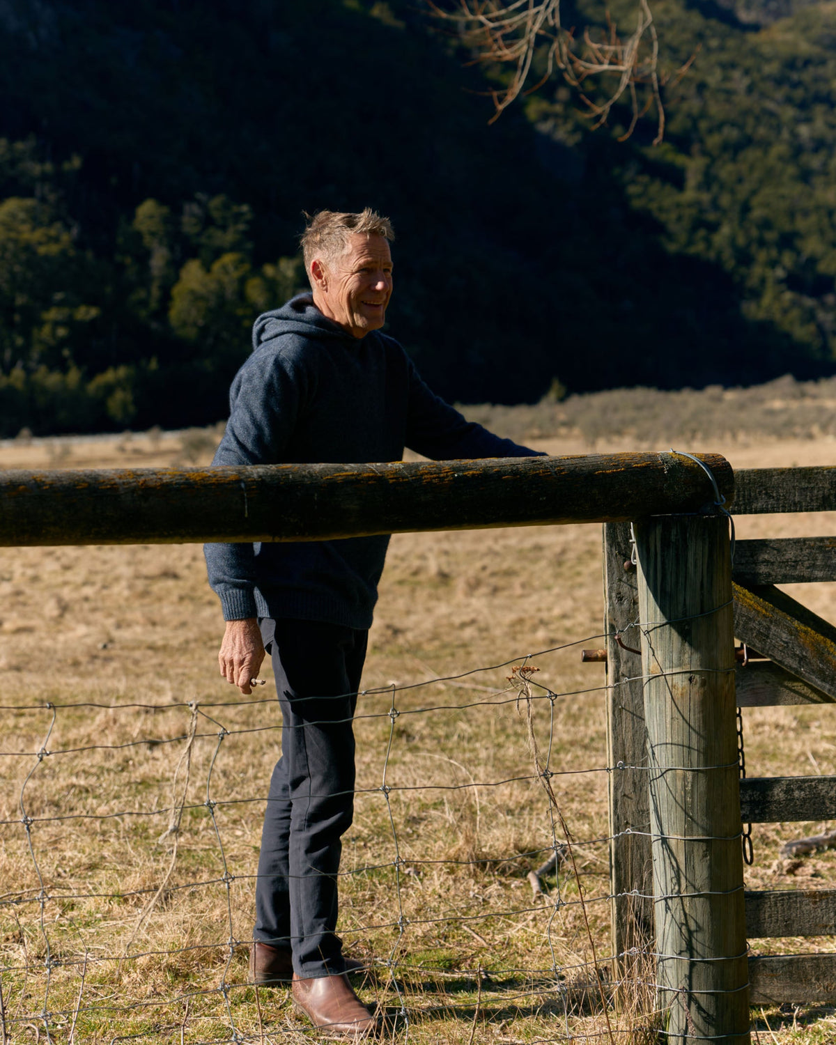 A man in a dark navy hoodie and dark pants stands outdoors beside a wooden fence in a dry grass field, with a forested mountain slope in the background.