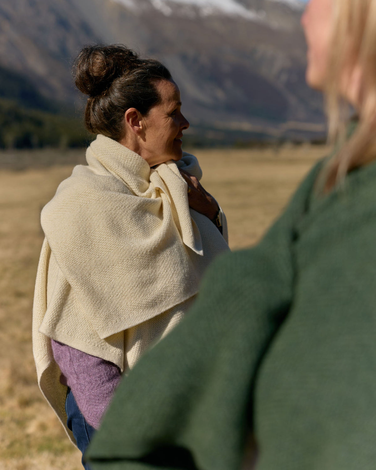 A woman outdoors in a mountain landscape, wrapped in a cream-colored knit blanket or shawl. Another person is partially visible in the foreground wearing a dark green top.