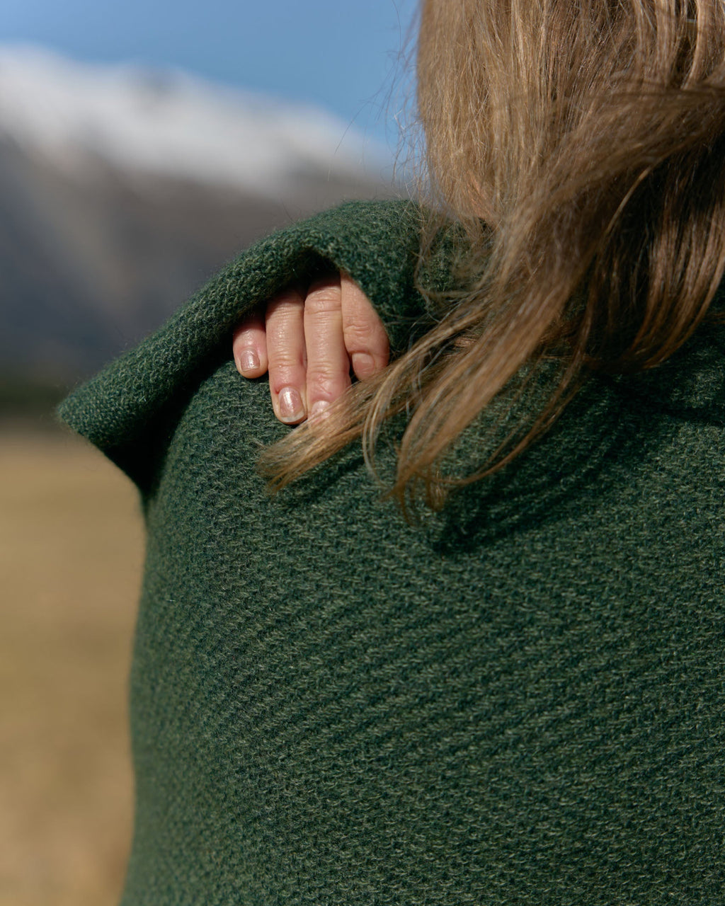 A close-up lifestyle shot of a person outdoors wearing a dark green textured knit wrap or blanket draped over their shoulder, with a blurred mountain landscape in the background.