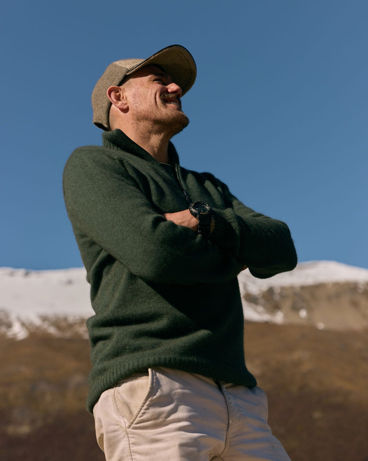 A man wearing a dark green knit sweater, beige pants, and a tan baseball cap stands outdoors with his arms crossed, looking upward against a clear blue sky and snowy mountains in the background.