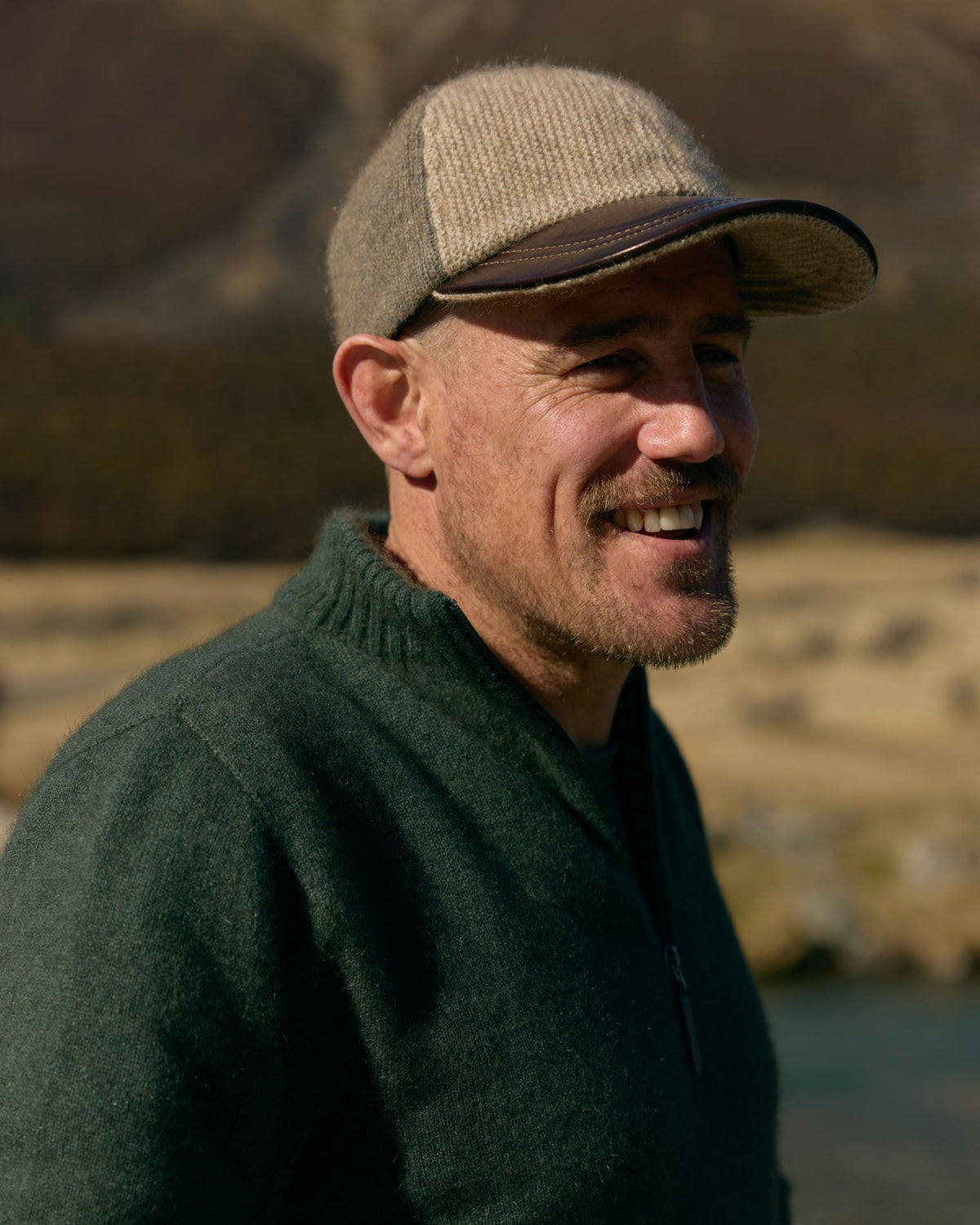 A smiling man wearing a beige tweed cap and a dark green knit quarter-zip sweater, photographed outdoors against a softly blurred natural background.