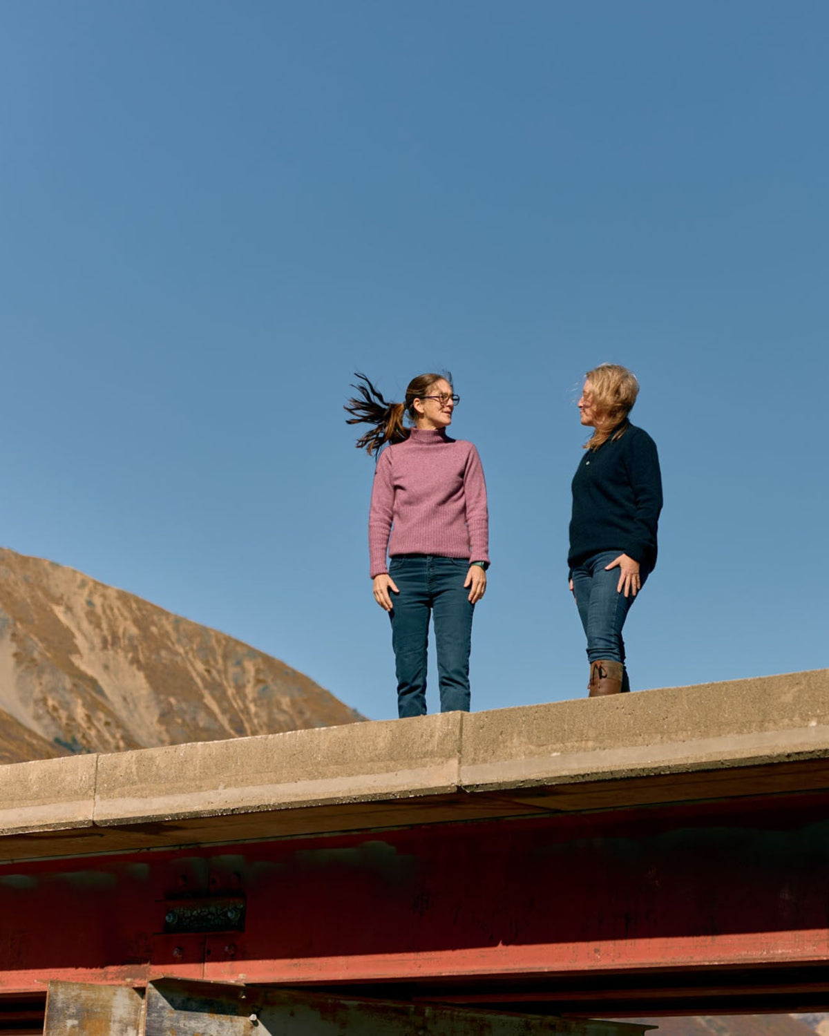 Two women standing on a concrete bridge overlooking a mountain under a clear blue sky, wearing casual outdoor clothing.
