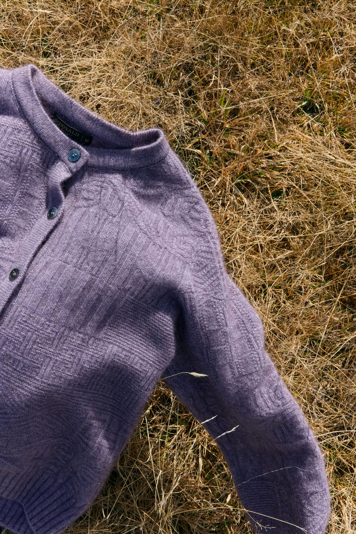 A close-up of a textured lavender knit cardigan laid on dry golden grass outdoors.