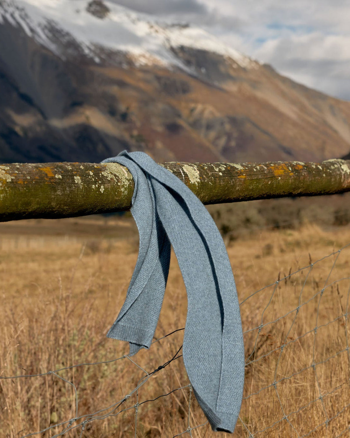 A light blue knit garment draped over a rustic wooden fence in an outdoor mountain landscape with dry grass and snow-capped peaks in the background.