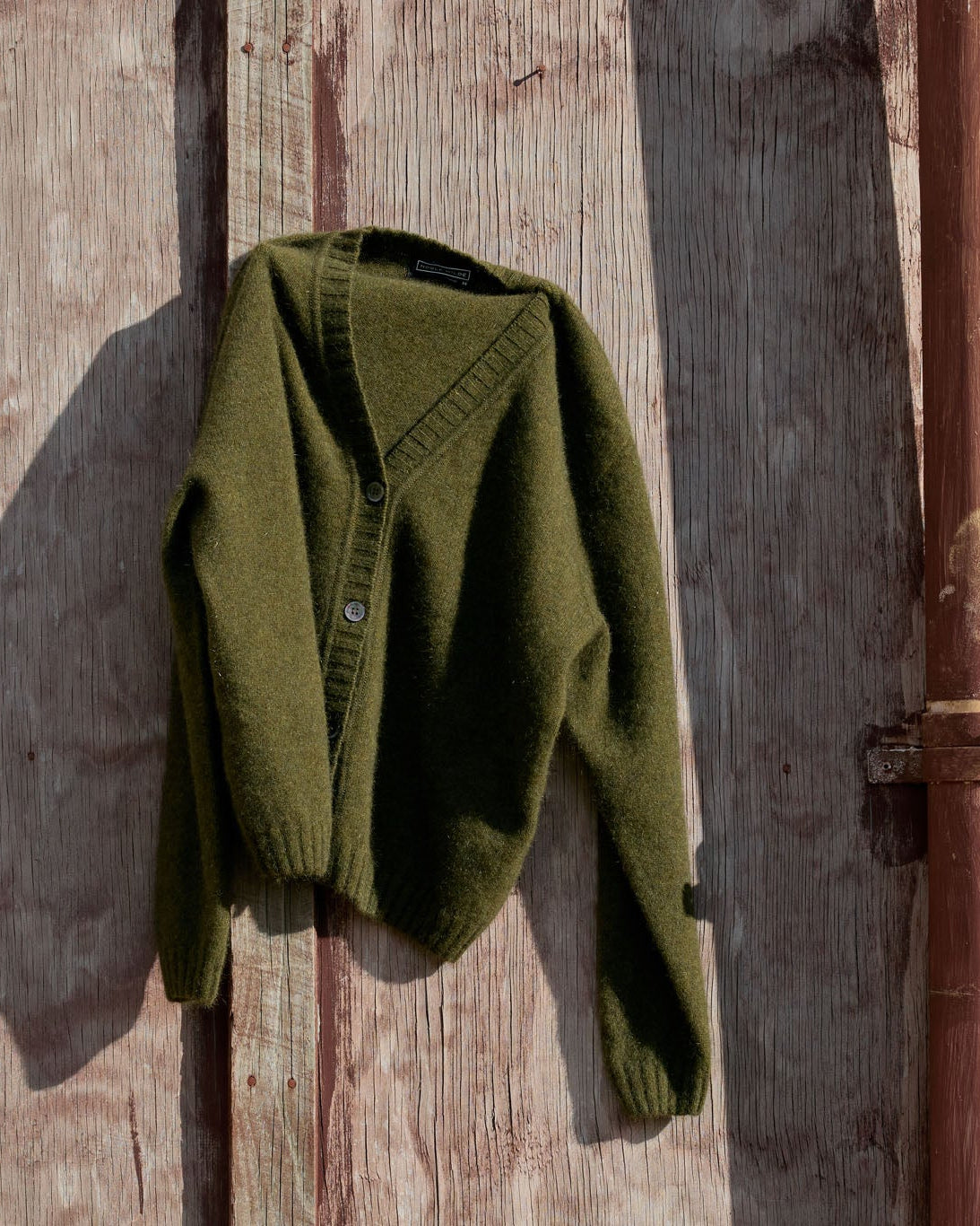 An olive green knit cardigan sweater hanging against a weathered wooden wall with a rusted metal pipe on the right.