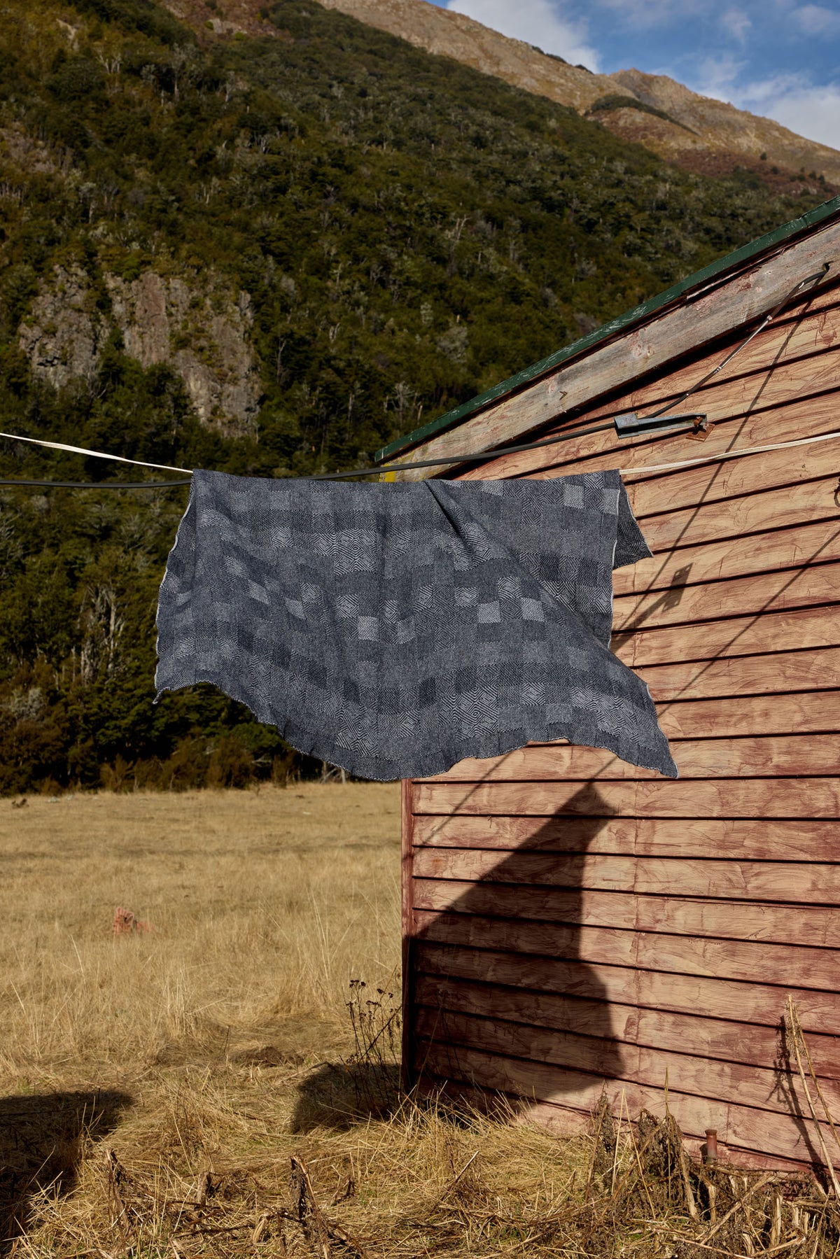 A dark gray patterned knit blanket or scarf hangs from a wire line outside a rustic wooden cabin, with a dry grass field and a forested mountain in the background.