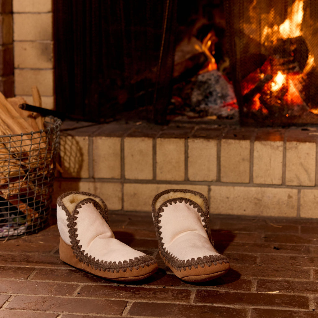 A pair of white moccasin-style slipper boots with brown braided trim and a tan sole, placed on a brick hearth in front of a lit fireplace.