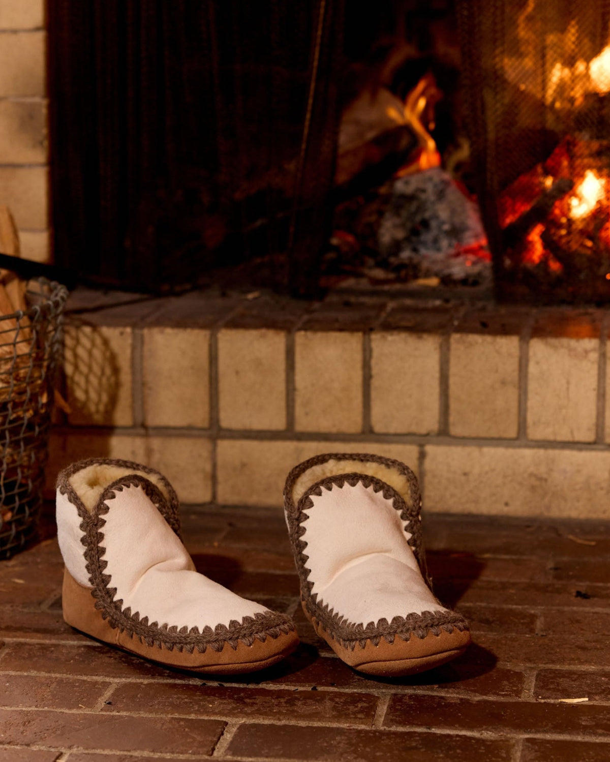 A pair of white moccasin-style slipper boots with brown braided trim and a tan sole, placed on a brick hearth in front of a lit fireplace.