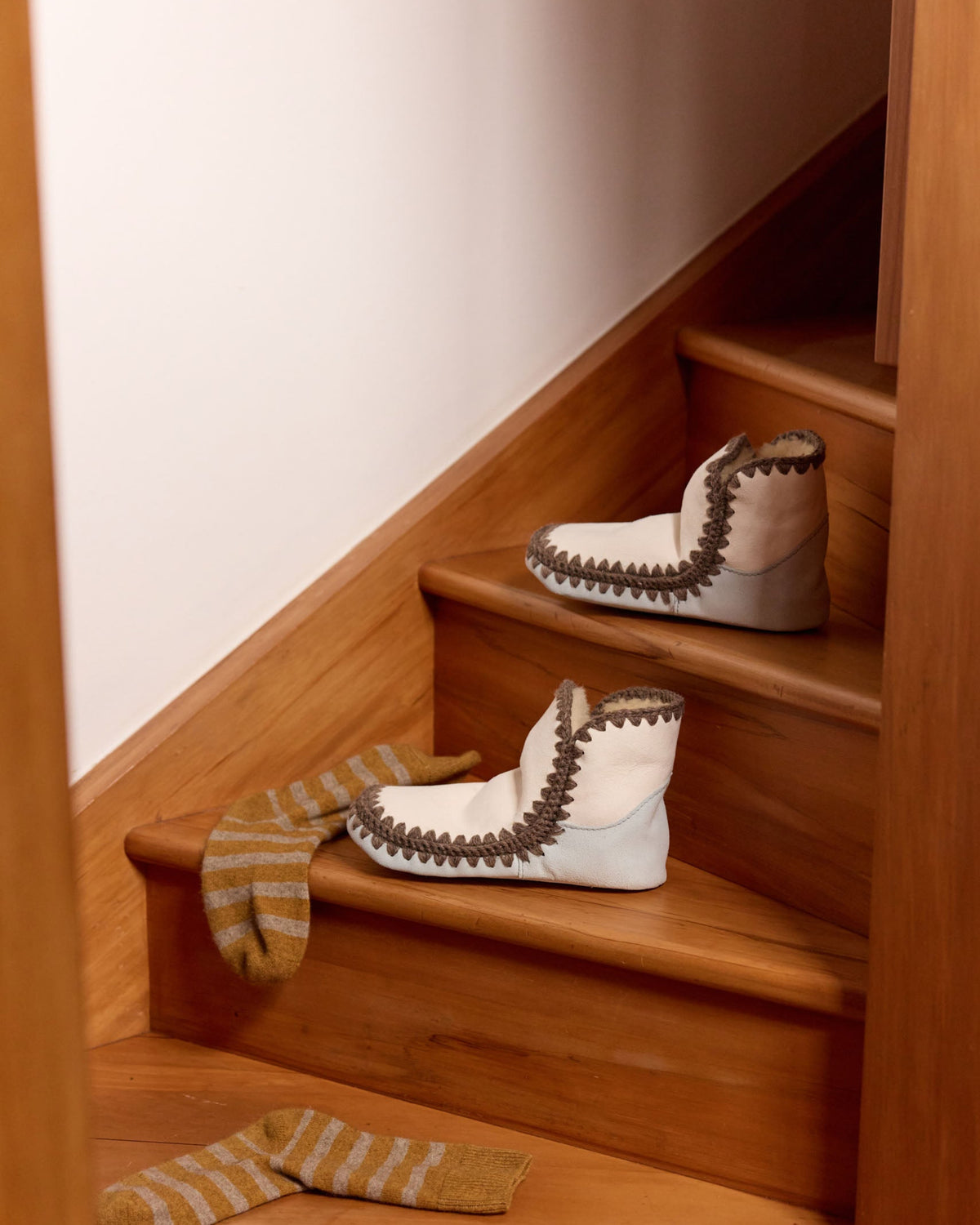 Three white moccasin-style boots with brown trim are arranged on wooden stairs, with matching striped socks placed on the steps below.