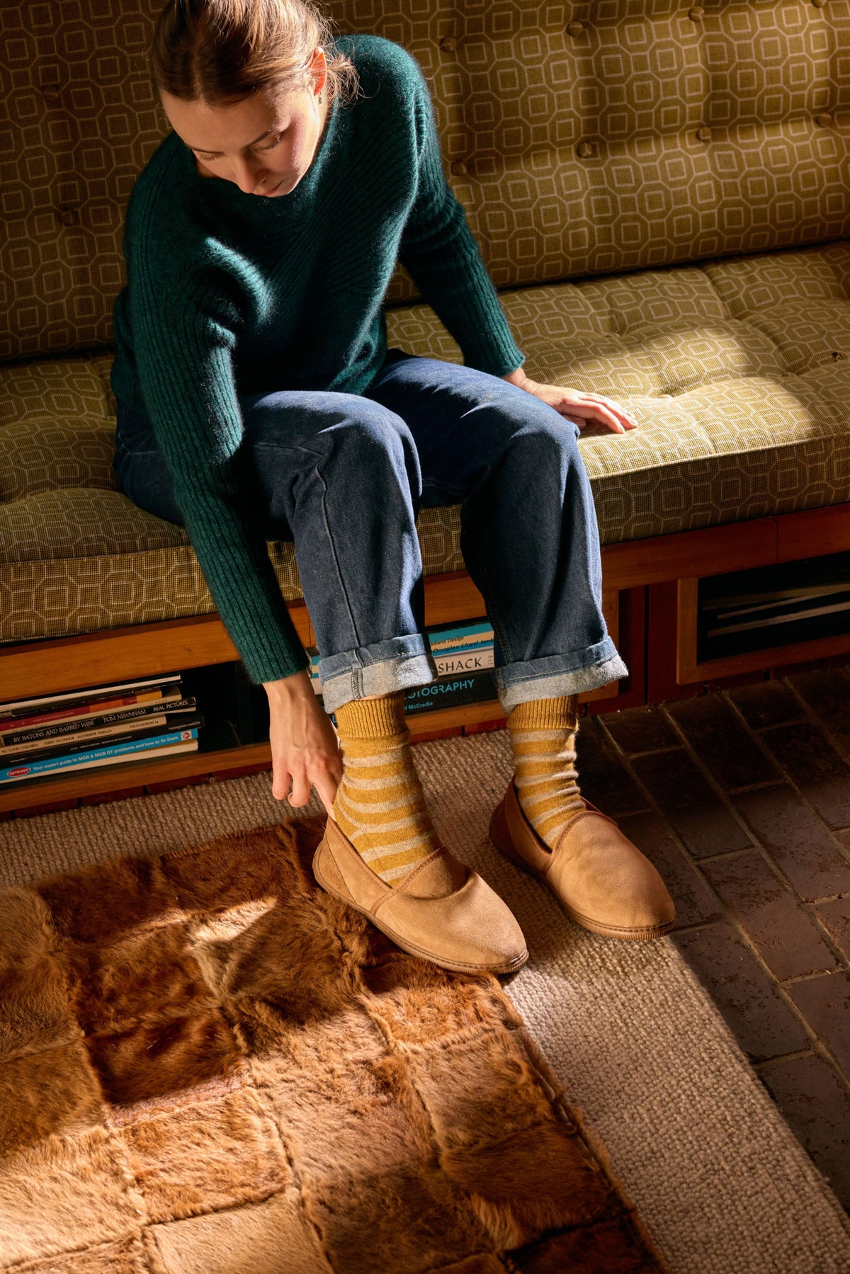 possum merino A woman sits on a patterned sofa in a warmly lit room, adjusting tan slip-on shoes with striped socks. She wears a dark green sweater and blue jeans, with a brown rug and wooden floor visible.