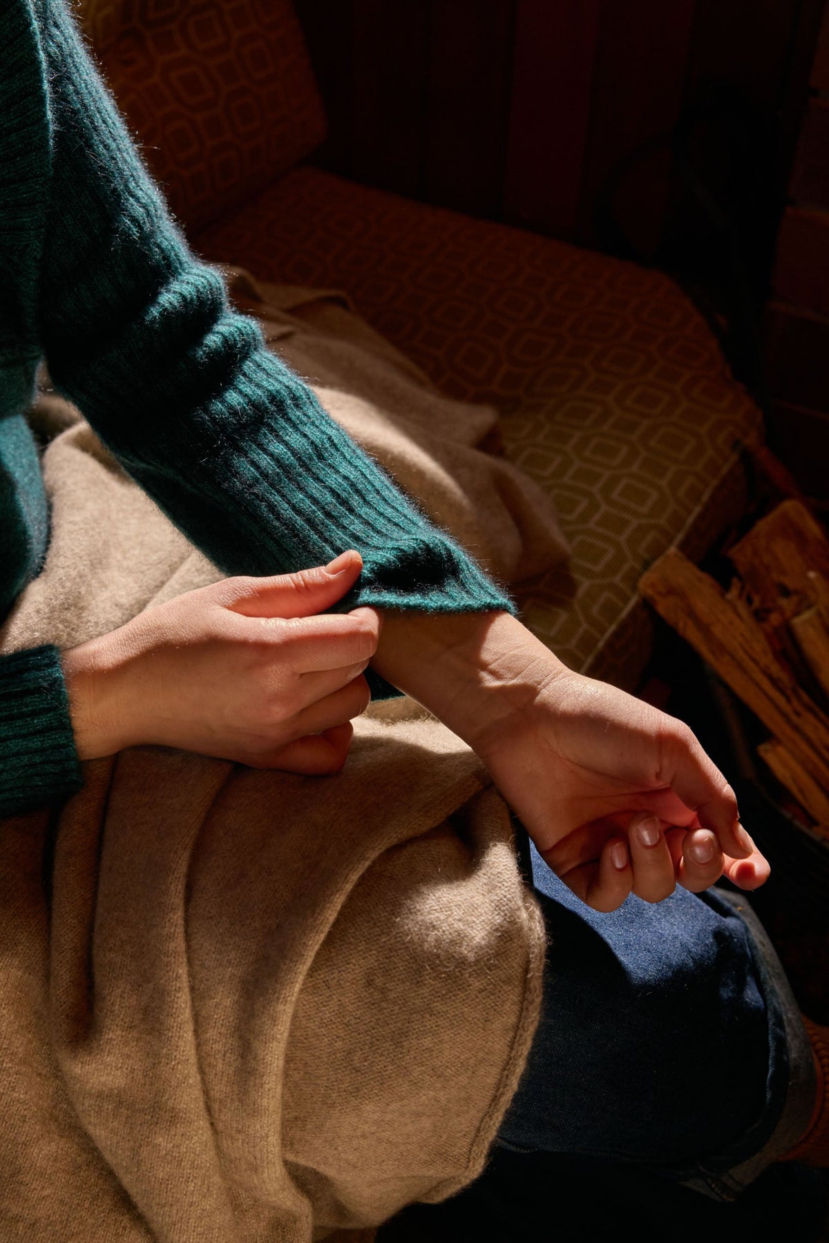 possum merino A close-up lifestyle shot of a person wearing a dark teal ribbed knit sweater and a beige cashmere wrap, sitting in warm, dimly lit surroundings.