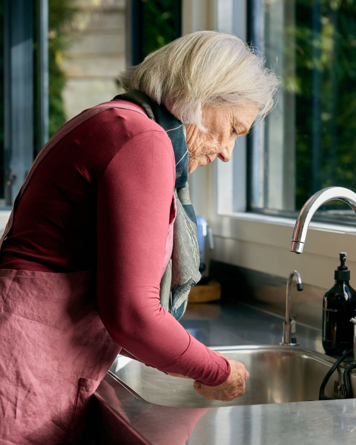 An older woman with white hair leans over a stainless steel kitchen sink, washing her hands. She is wearing a dusty rose long-sleeve top and a dark apron.