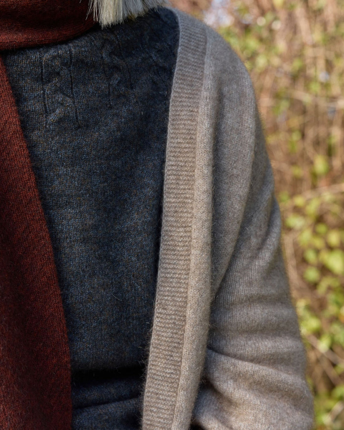 A close-up of a person wearing a beige knit cardigan, a dark gray knit sweater, and a rust-brown scarf, with a blurred outdoor background of dry grass and greenery.