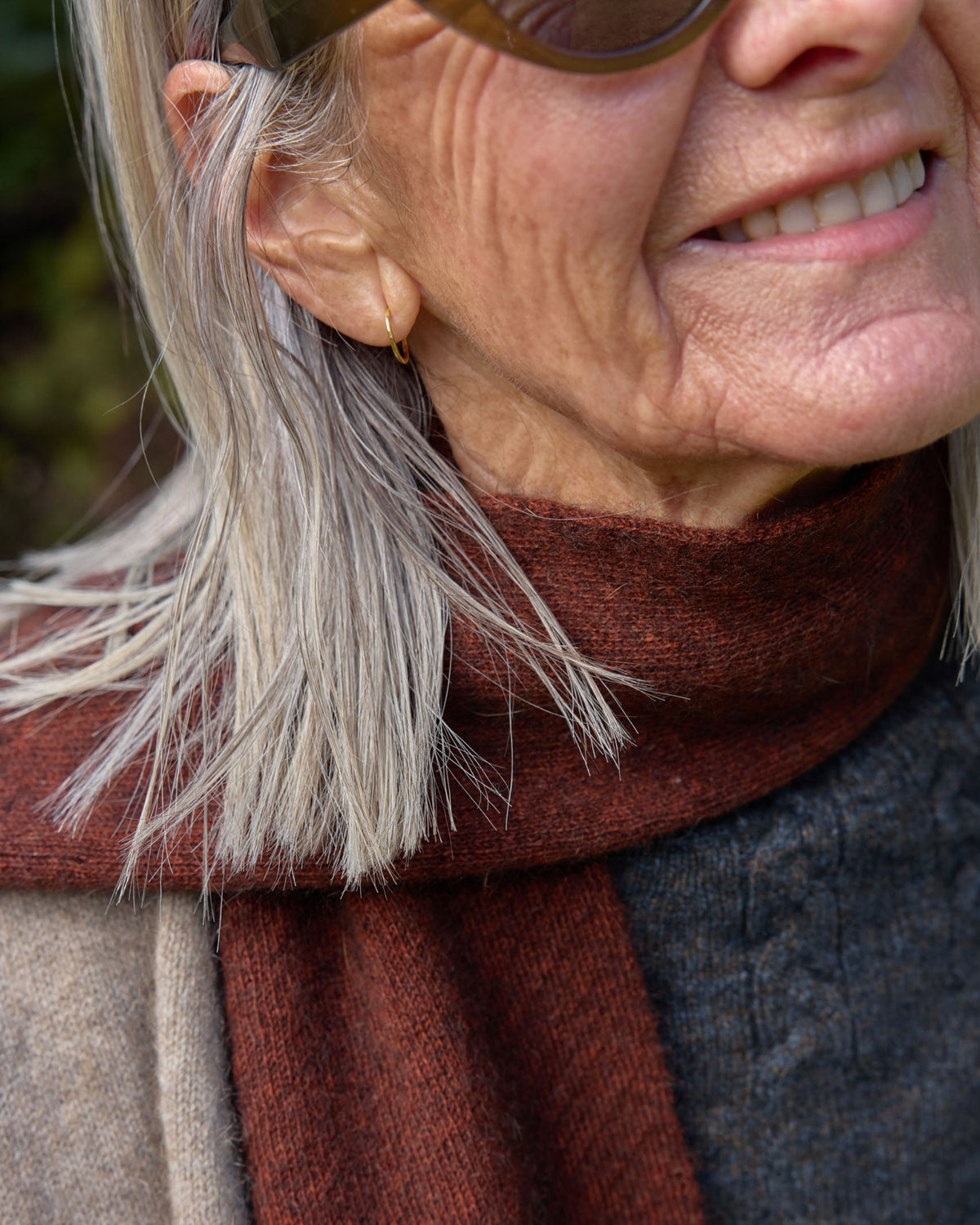 A close-up lifestyle shot of a smiling older woman wearing a dark rust-brown knit scarf, a gray knit sweater, and a beige coat.