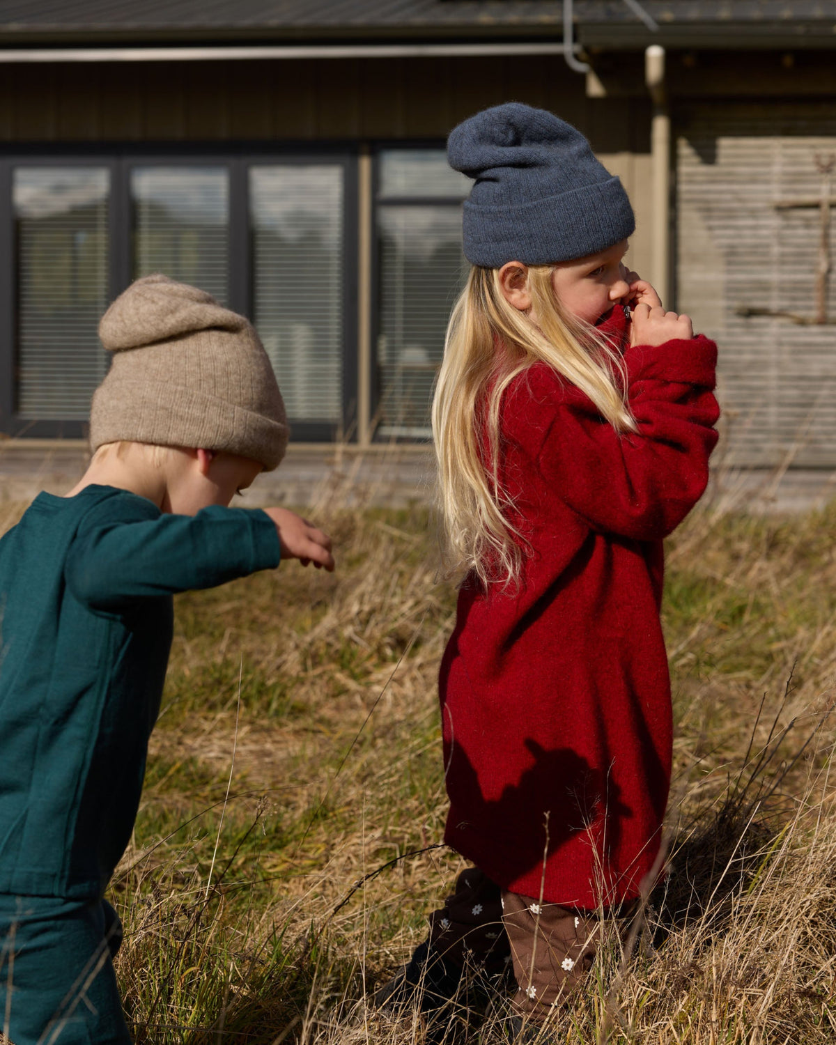 Two children outdoors in a grassy field in front of a house. The child on the right wears a dark blue knit beanie and a red knit sweater, while the child on the left wears a beige knit beanie and a teal knit outfit.