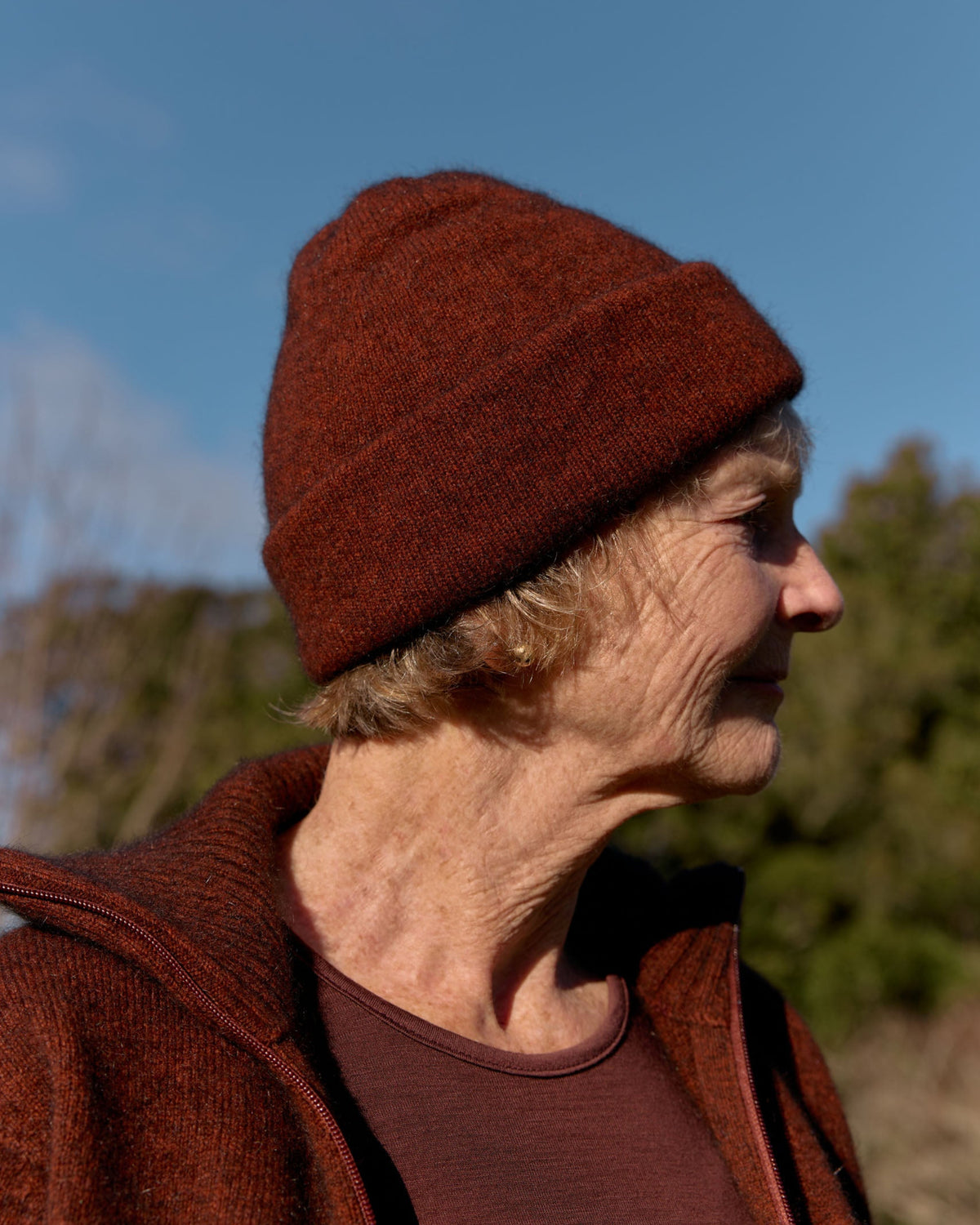 A person in profile wearing a textured rust-brown knit beanie and matching knit top outdoors against a blue sky and blurred trees.