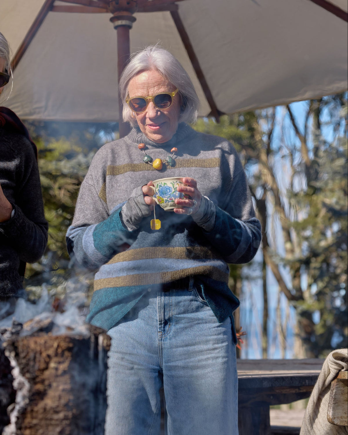 Two women standing outdoors under a large patio umbrella, each holding a small cup. The woman in the center wears a gray and teal striped sweater, blue jeans, and sunglasses, while the woman on the left wears a dark sweater and sunglasses.