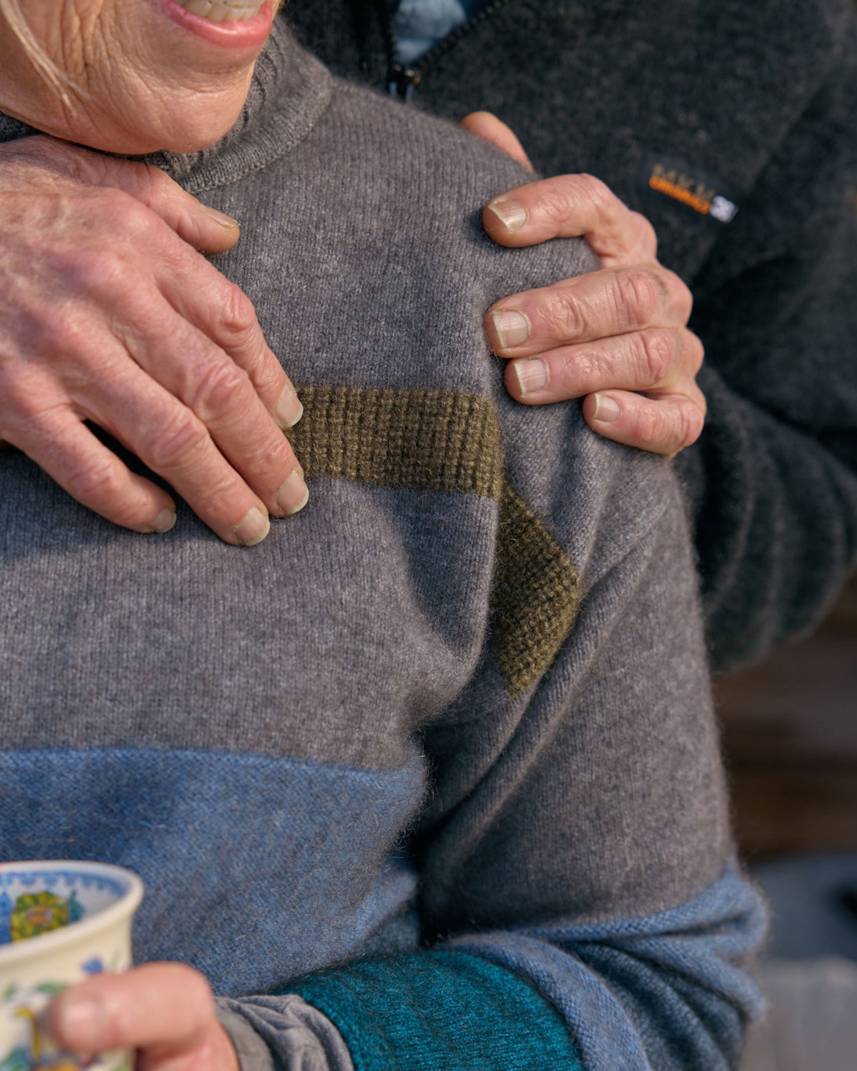 A close-up lifestyle shot of a person wearing a gray knit sweater with a contrasting olive green chest panel and teal ribbed cuffs, with another person's hands resting on their shoulders.