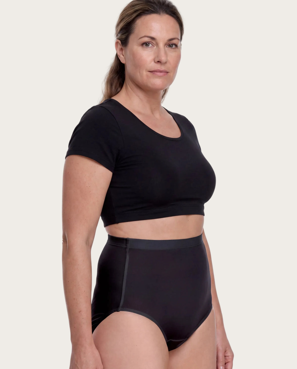 A woman modeling a black short-sleeve crop top and matching high-waisted black briefs against a plain white studio background.