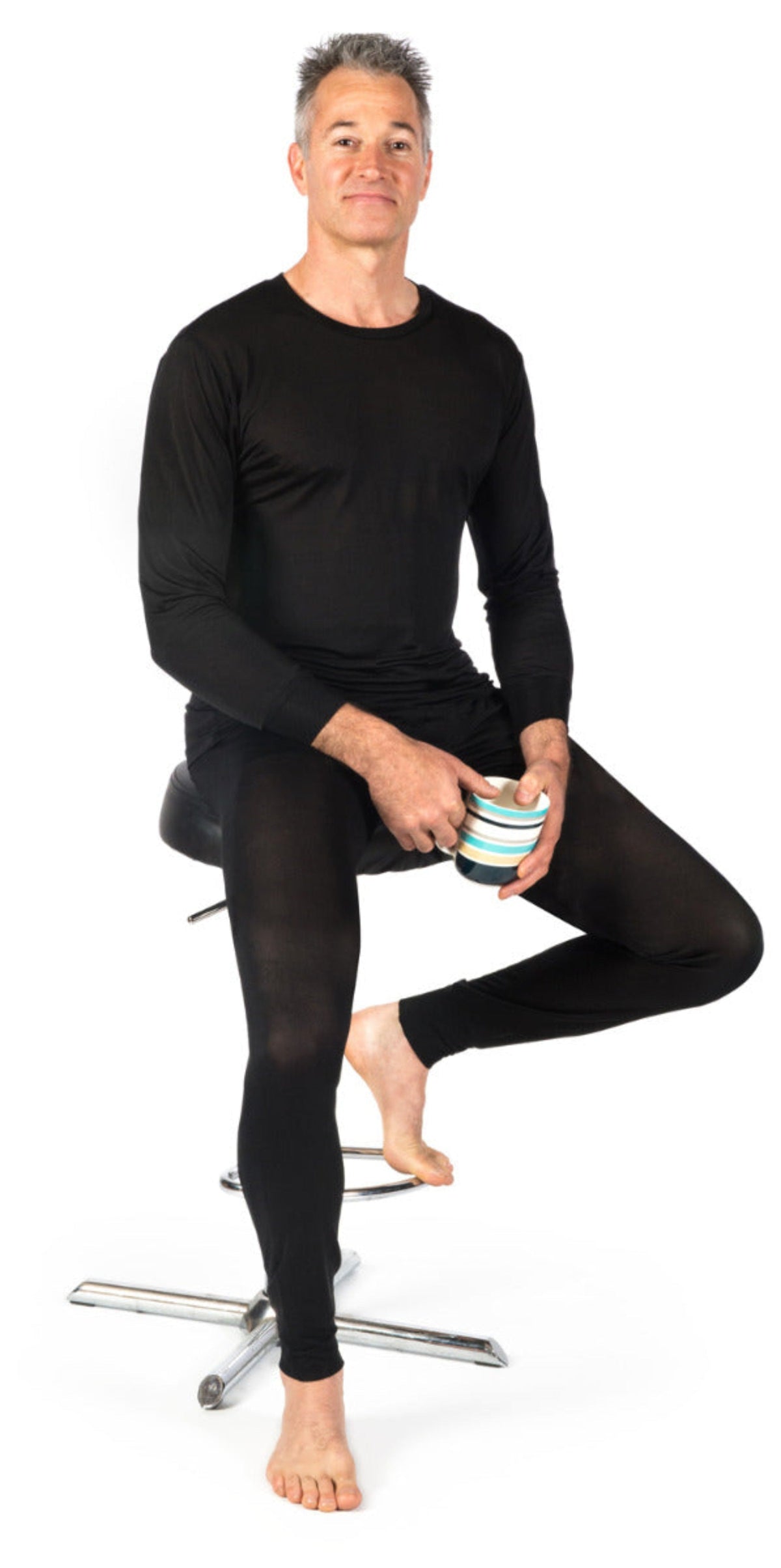 Man wearing silk black long-sleeve thermal underwear  and leggings sitting on a stool holding a striped mug.